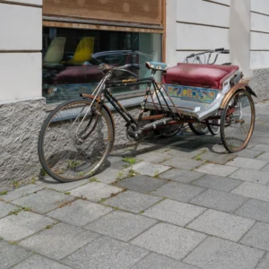 Eine traditionelle Rikscha mit drei Rädern, vorne ein Fahrrad und hinten ein dekorierter Fahrgastbereich, am Gehwegrand geparkt.