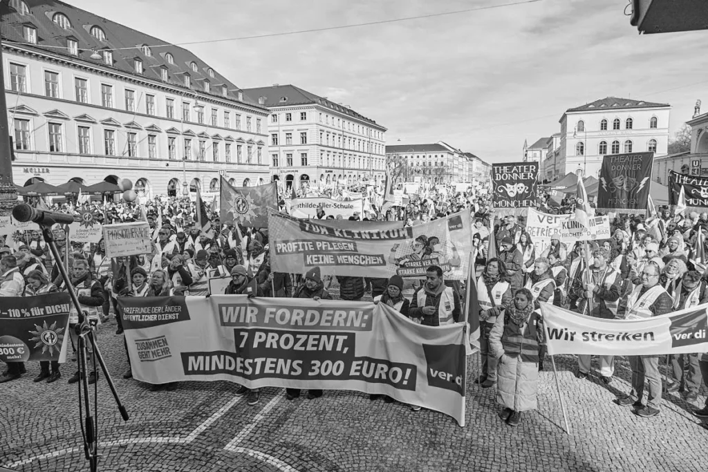 Münih Odeonsplatz'da gerçekleşen bir Ver.di Sendikasının Uyarı Grevi Göstertisi, "WIR FORDERN" (Talep Ediyoruz) yazılı pankartlar taşıyan işçiler ve çevredeki klasik binalar, siyah beyaz çekim.