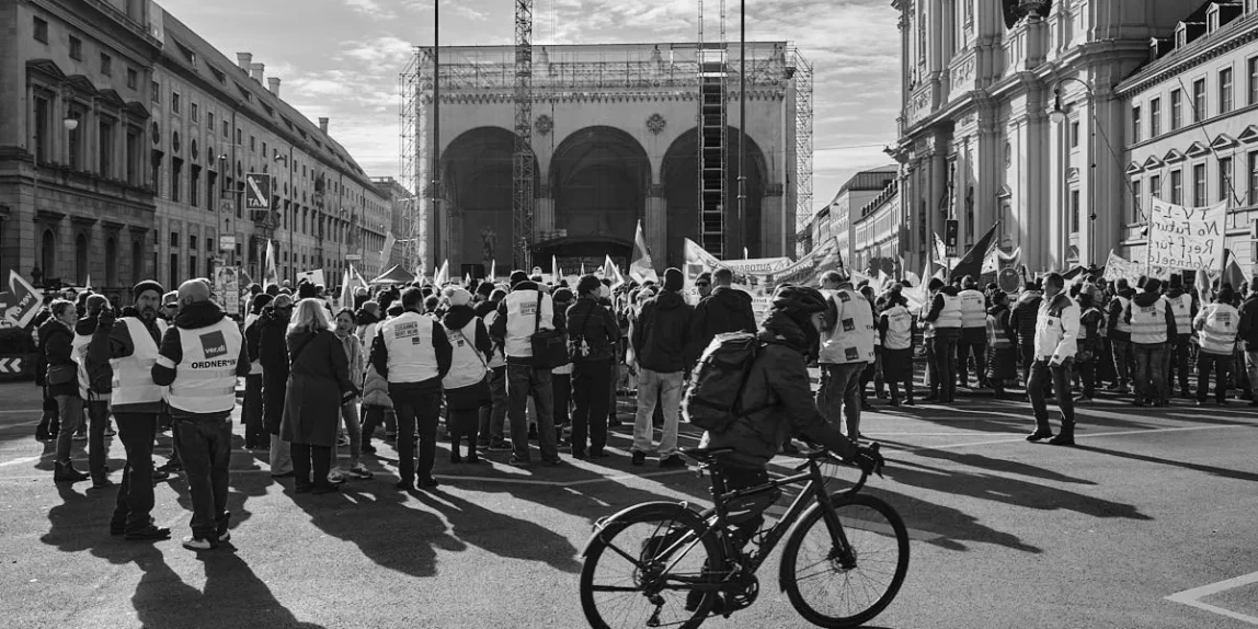 Münih Odeonsplatz'da, ön planda bisikletli bir kişinin gölgesi, arkada protesto pankartları ve inşaat halindeki Feldherrnhalle binası, siyah beyaz çekim.