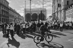 Münih Odeonsplatz'da, ön planda bisikletli bir kişinin gölgesi, arkada protesto pankartları ve inşaat halindeki Feldherrnhalle binası, siyah beyaz çekim.