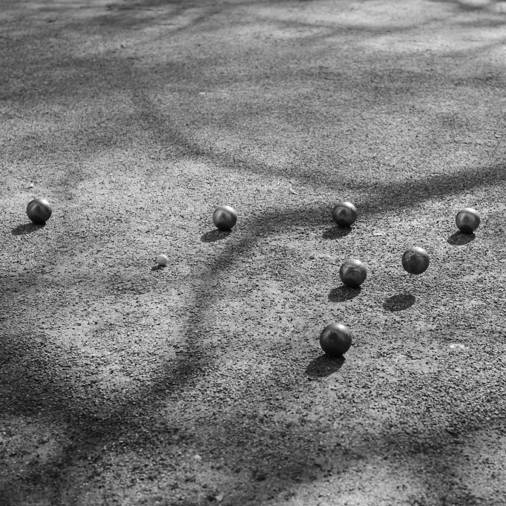 Siyah beyaz, üstten çekim perspektifiyle Hofgarten daToprak zemindeki petank topları ve üzerlerine düşen ağaç gölgeleri.