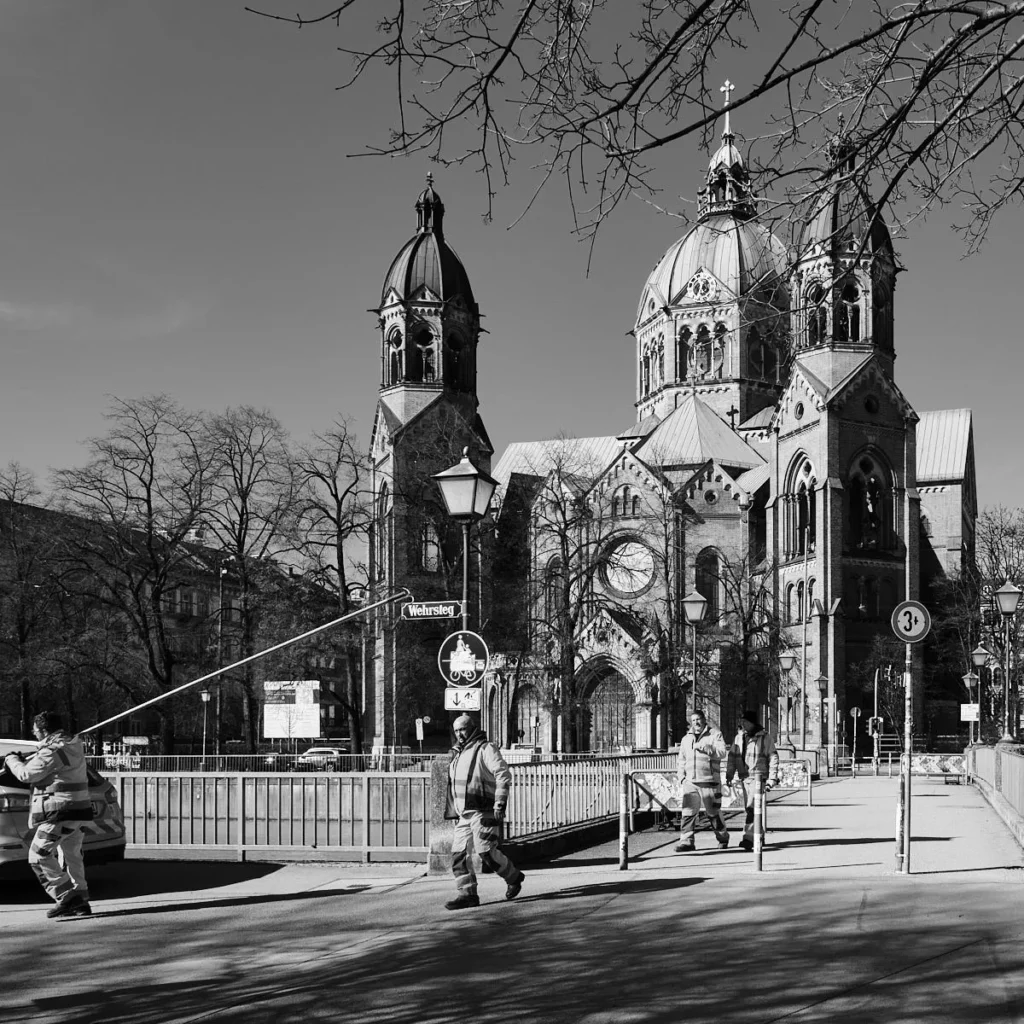 Arka planda tarihi bir kilisenin (St. Lukas) kubbeli kulelerinin göründüğü, ön planda ise iş kıyafetleri içindeki insanların köprüden geçtiği siyah beyaz şehir manzarası.