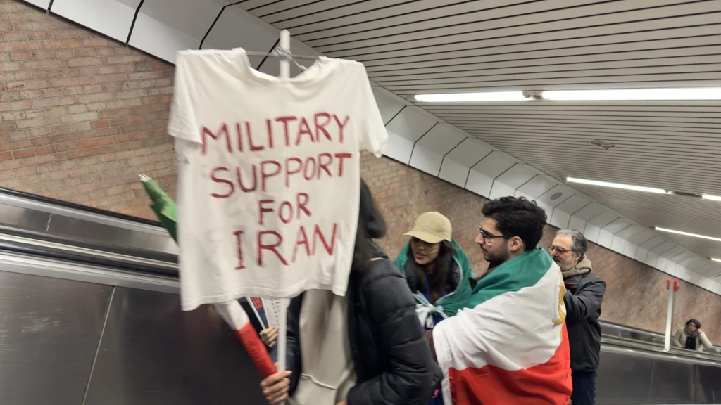 Theresienwiese’deki İran protestosuna giderken Münih U-Bahn’da “Military Support for Iran” yazılı pankart taşıyan göstericiler.