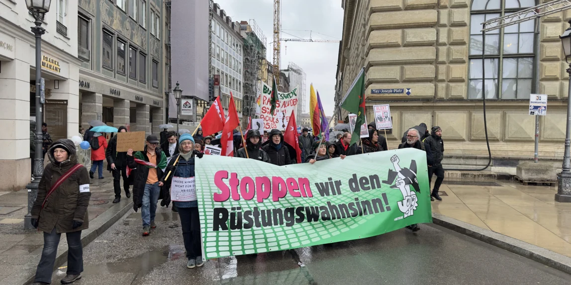Münih caddelerinde, üzerinde Almanca "Stoppen wir den Rüstungswahnsinn!" (Silahlanma çılgınlığını durduralım) yazılı büyük yeşil bir pankart taşıyan protestocu grubu ve çevredeki güvenlik önlemleri,