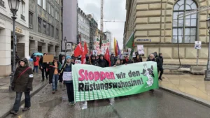 Münih caddelerinde, üzerinde Almanca "Stoppen wir den Rüstungswahnsinn!" (Silahlanma çılgınlığını durduralım) yazılı büyük yeşil bir pankart taşıyan protestocu grubu ve çevredeki güvenlik önlemleri,
