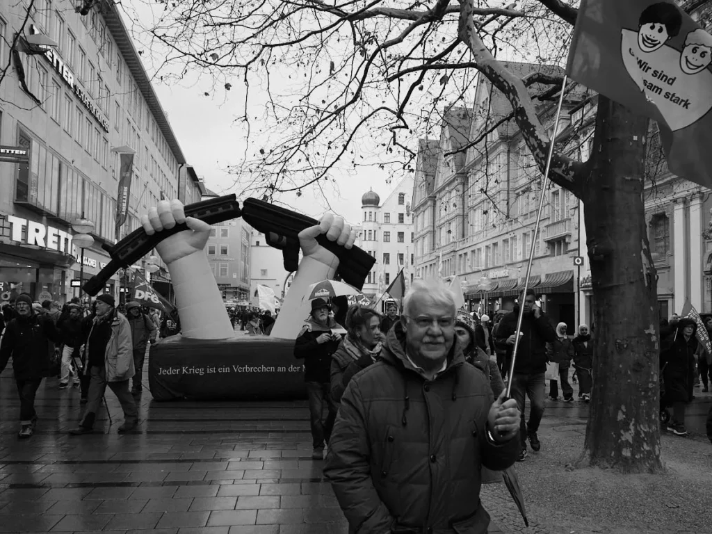 Siyah beyaz karede, arka planda "Jeder Krieg ist ein Verbrechen" (Her savaş bir suçtur) yazılı şişme bir enstalasyon ve ön planda gülümseyen yaşlı bir adam ile bayrak taşıyan kalabalık.