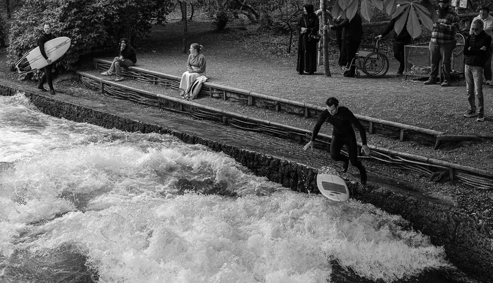 Zuschauer und Surfer an der legalisierten Eisbachwelle in München: Ein Surfer springt vor Publikum mit seinem Board in den Fluss.