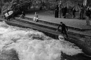 Zuschauer und Surfer an der legalisierten Eisbachwelle in München: Ein Surfer springt vor Publikum mit seinem Board in den Fluss.